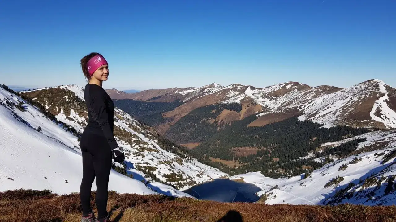 Vue panoramique depuis le pic du lion, une femme souriante admire le paysage montagneux enneigé et un lac gelé.