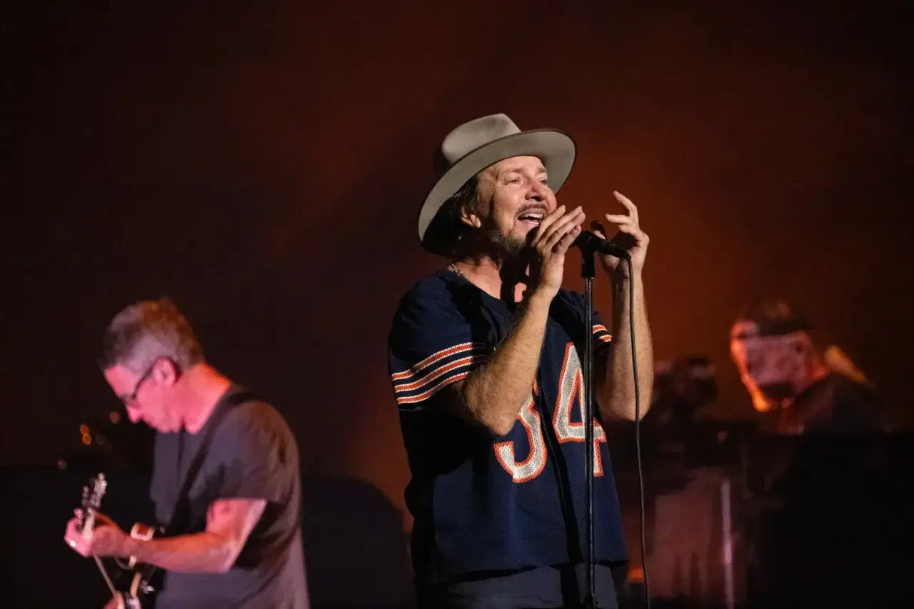 Eddie Vedder, wokalista Pearl Jam, w kapeluszu i koszulce z numerem 34, śpiewa do mikrofonu na koncercie.