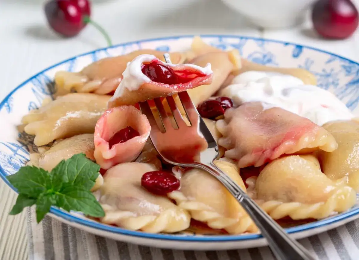 Pyszne pierogi z czereśniami, polane śmietaną, z listkiem mięty. Widok na talerzu, jeden pieróg na widelcu.