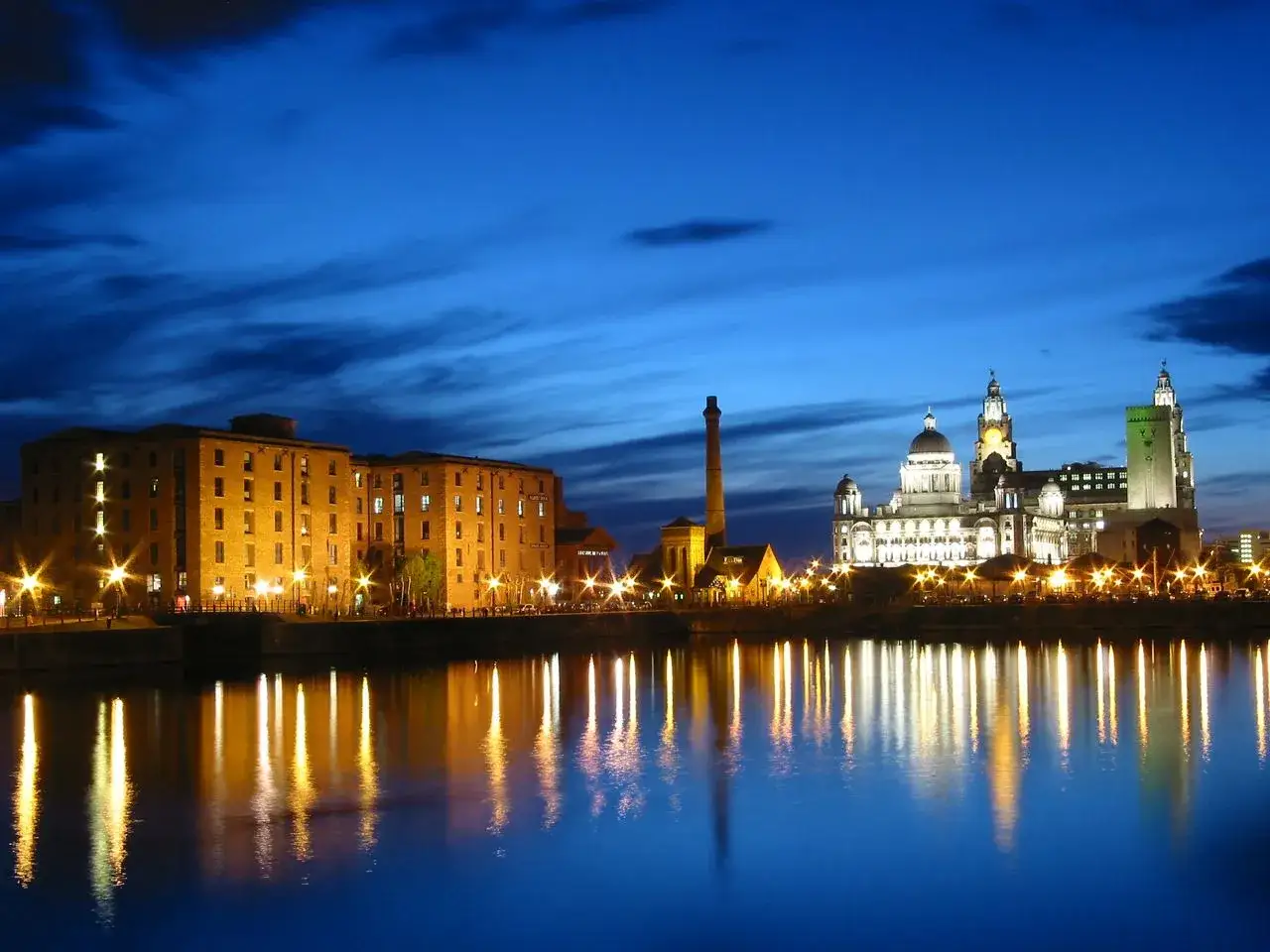 La nuit, les docks illuminés et les bâtiments emblématiques de Liverpool se reflètent dans l'eau. Une belle image pour planifier votre visite de Liverpool en 1 jour.