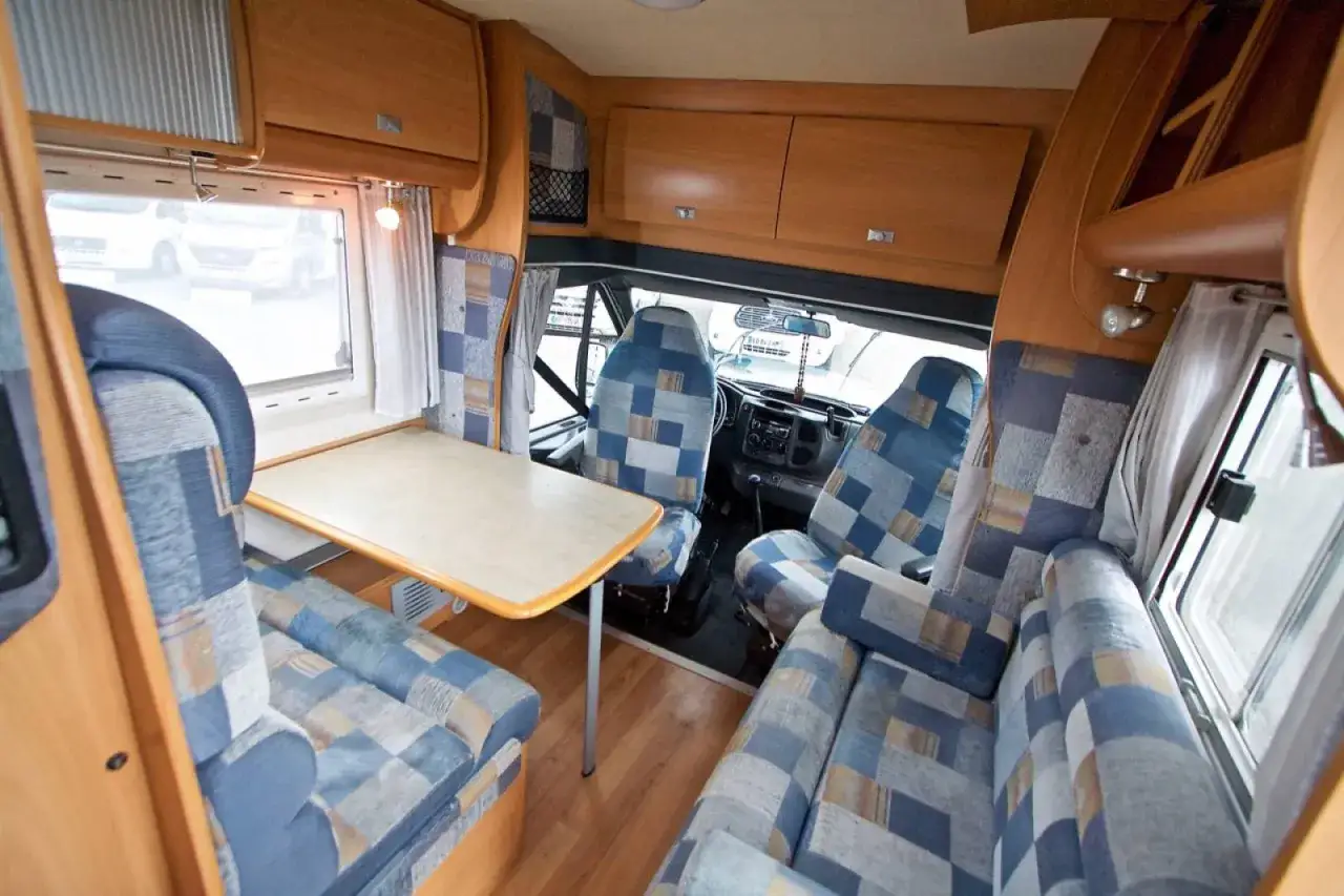 Int&eacute;rieur d'un camping-car avec si&egrave;ges bleus &agrave; carreaux, table et cabine de pilotage. Un espace confortable pour un voyage genesis 30.