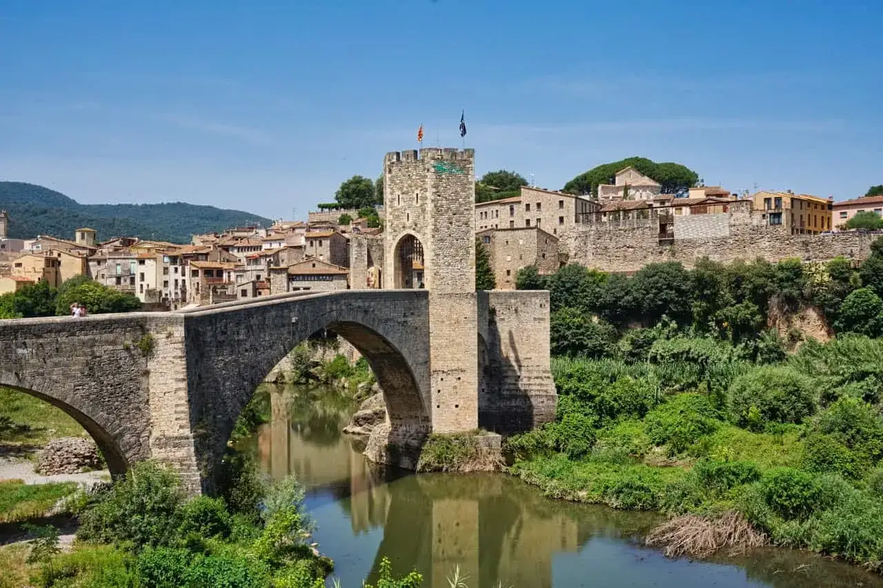 Mittelalterliche Br&uuml;cke mit Turm in Besal&uacute;, Spanien. Die steinerne Br&uuml;cke &uuml;berspannt einen Fluss, dahinter erstreckt sich die historische Stadt mit ihren H&auml;usern und Mauern.
