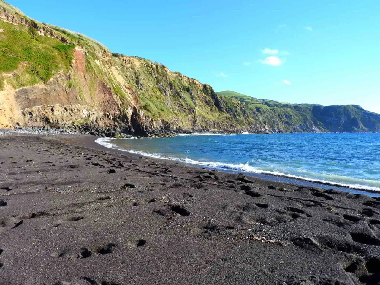 Schwarzer Sandstrand mit Fußspuren, steile, grüne Klippen und das blaue Meer – ein malerischer azoren strand.