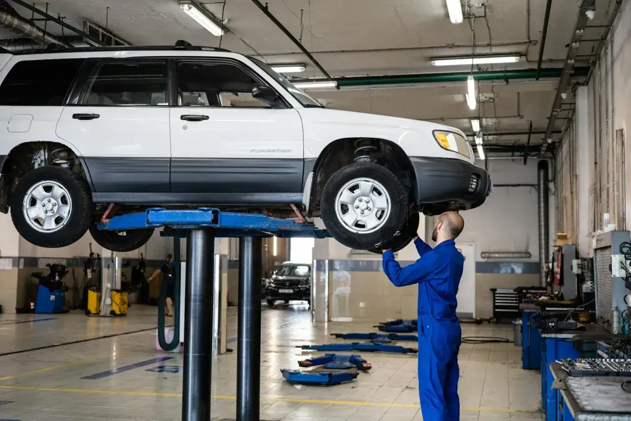 Mechanik w niebieskim kombinezonie pracuje przy podniesionym samochodzie. W tle widać narzędzia i inne samochody, a także klocki ceramiczne na podłodze.