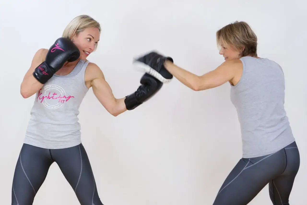 Two women in athletic wear practice boxing. One woman throws an uppercut to the body, her black glove aimed at her partner's mitt.