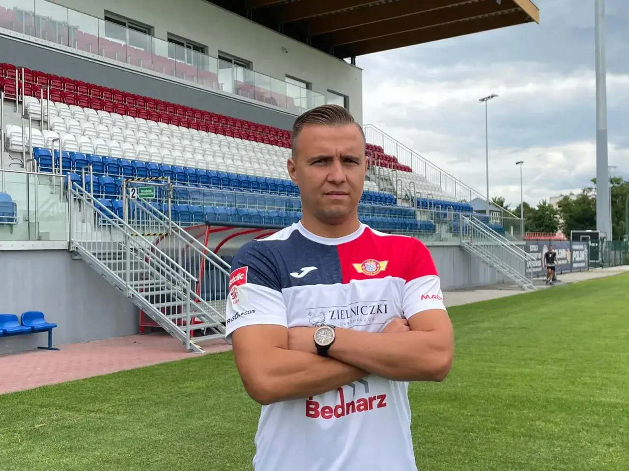 Michał Kaszuba w stroju sportowym na tle stadionu z czerwono-biało-niebieskimi trybunami.