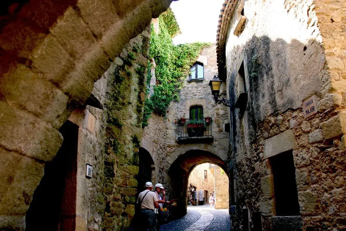 Eine schmale Gasse in Pals, Spanien, mit alten Steinh&auml;usern, Efeu und einem Torbogen.