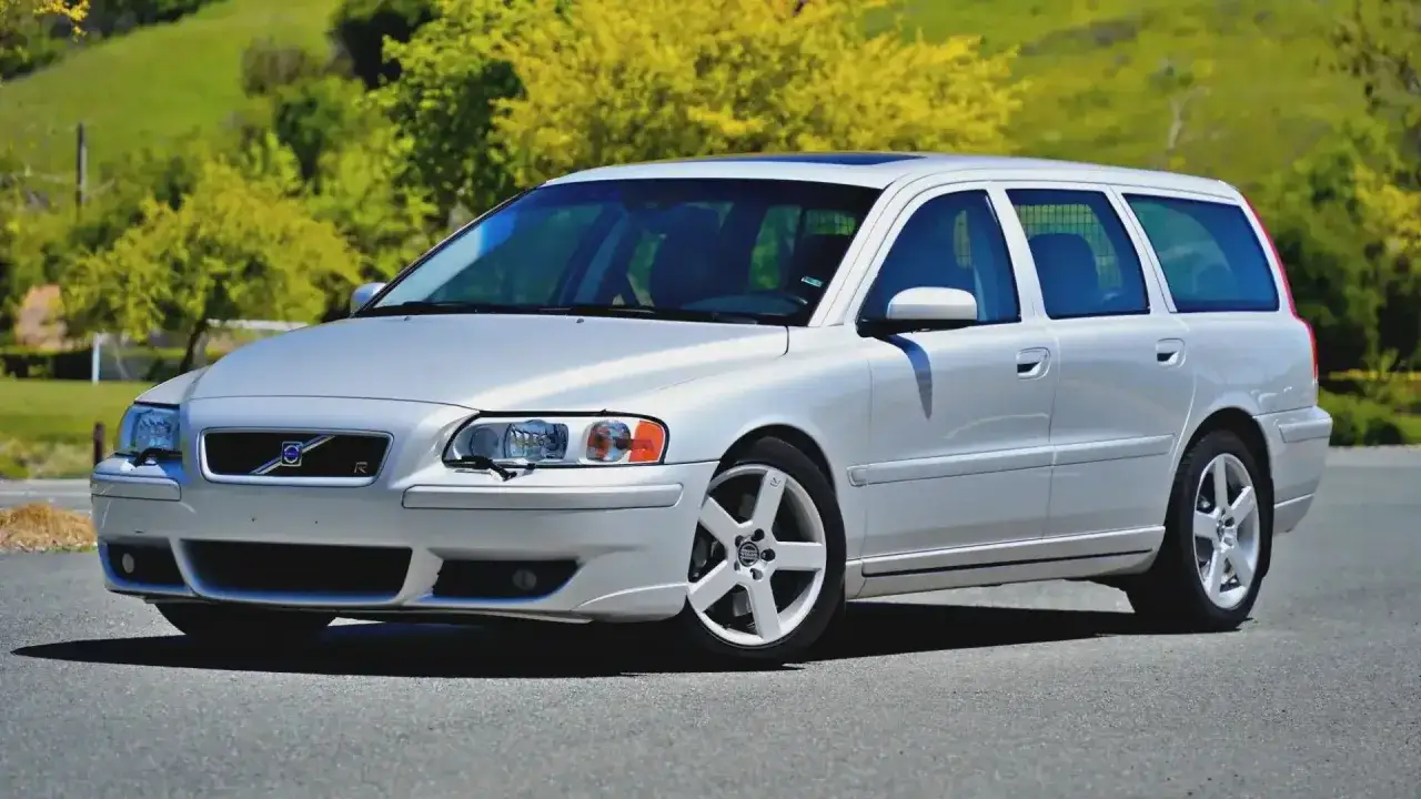 Srebrne Volvo V70 R, klasyczny sleeper car, zaparkowane na tle zielonych wzg&oacute;rz i ż&oacute;łtych drzew.