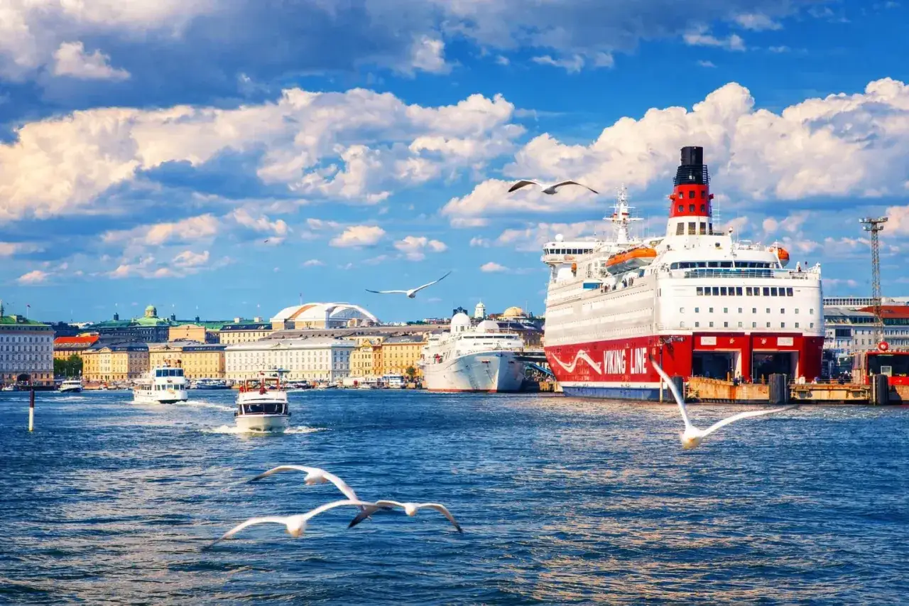 Duży prom Viking Line cumuje w porcie, w tle widać panoramę Helsinek. Mewy szybują nad wodą.