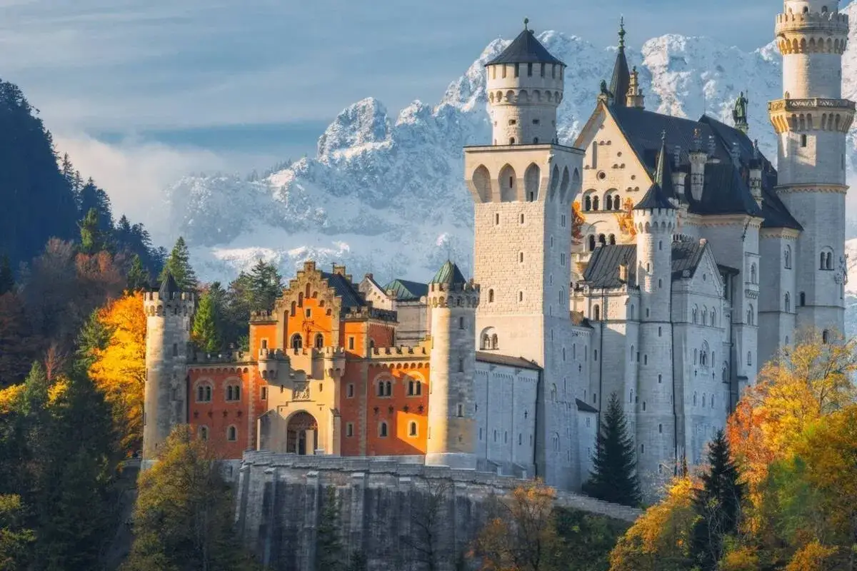 El castillo del rey loco, un sueño de piedra y torres, se alza majestuoso entre montañas nevadas y bosques otoñales.