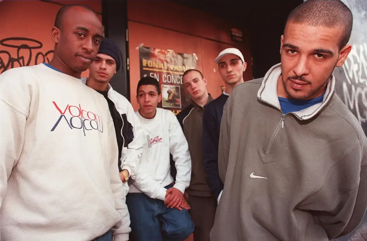 Un groupe de jeunes hommes, dont un ancien rappeur fran&ccedil;ais, pose devant un mur orange.