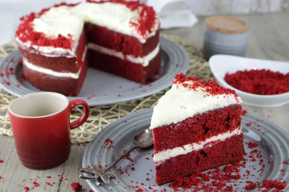 Una rebanada de tarta red velvet con glaseado blanco y migas rojas, junto a una taza roja y el resto del pastel.