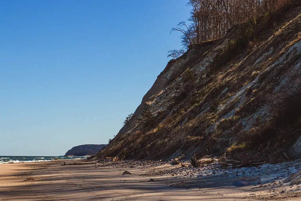 Plaża i klif w Wolińskim Parku Narodowym. Słońce oświetla zbocze porośnięte trawami i drzewami.