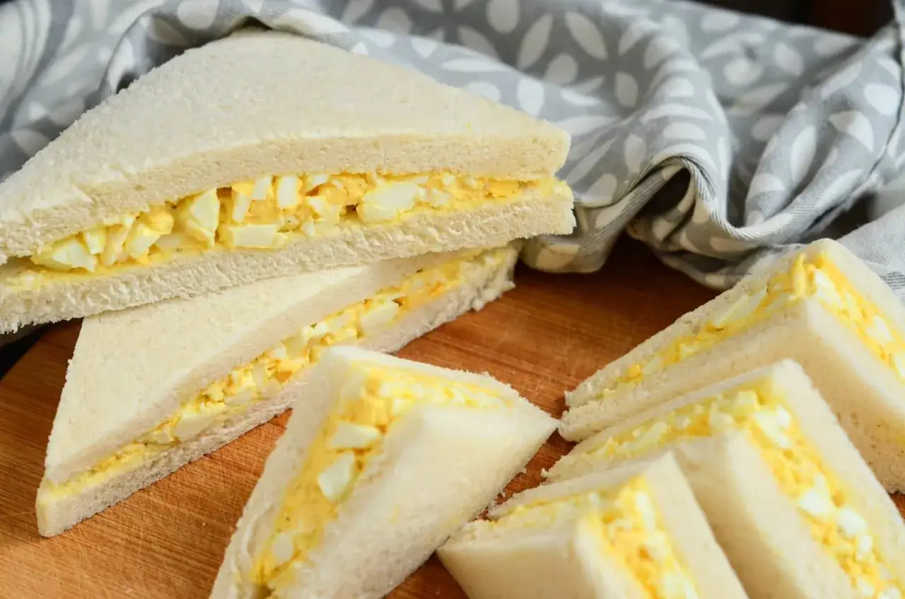 Plusieurs sandwichs aux &oelig;ufs coup&eacute;s en triangles, sur une planche en bois, avec un torchon &agrave; motifs.
