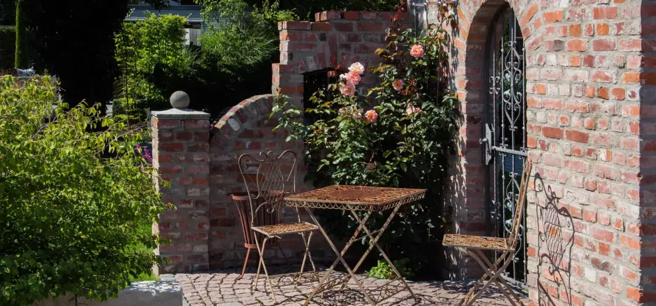 Ein romantischer Garten mit einer alten Ziegelsteinmauer und einem schmiedeeisernen Tischset. Rosen ranken an der natursteinmauer garten empor.