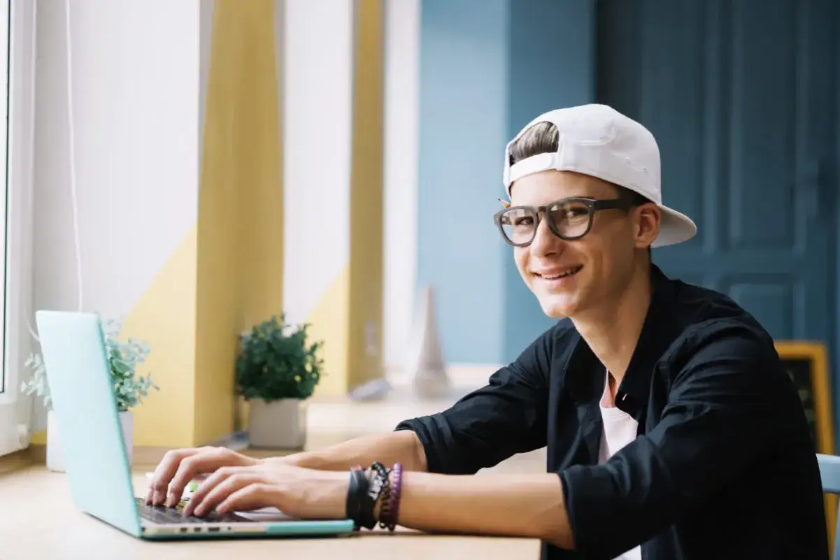 Uśmiechnięty nastolatek w czapce i okularach pracuje na laptopie. Zastanawia się, od ilu lat można pracować, by zarobić na taki sprzęt.