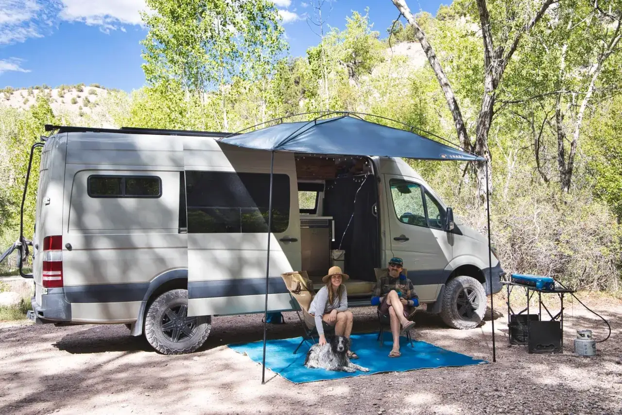 Ein Paar mit Hund entspannt vor einem Wohnmobil. Die Markise spendet Schatten. Perfekt für die Wohnmobil Packliste!