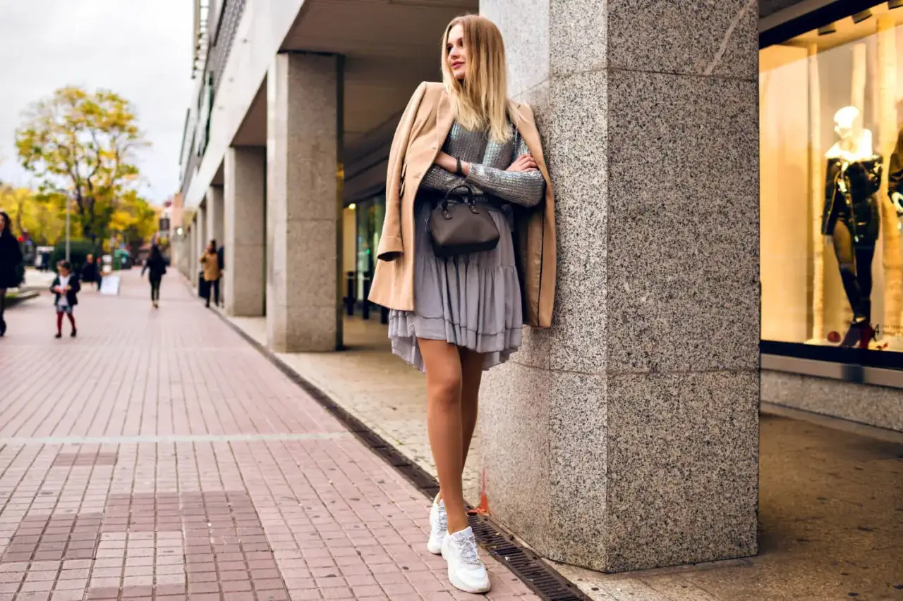 Stylowa kobieta w beżowym płaszczu i szarej sukience, zastanawia się jakie buty do sukienki będą najlepsze. Postawiła na białe sneakersy.