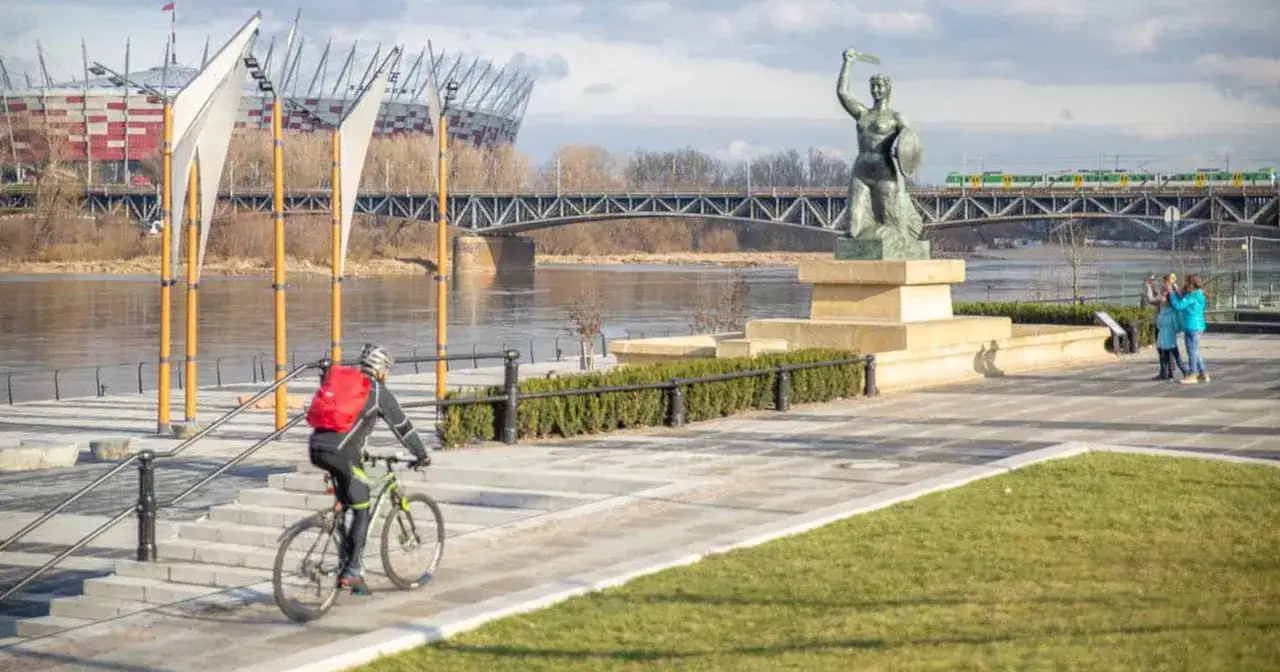 Rowerzysta na bulwarach wiślanych w Warszawie, w tle Syrenka, most i Stadion Narodowy.