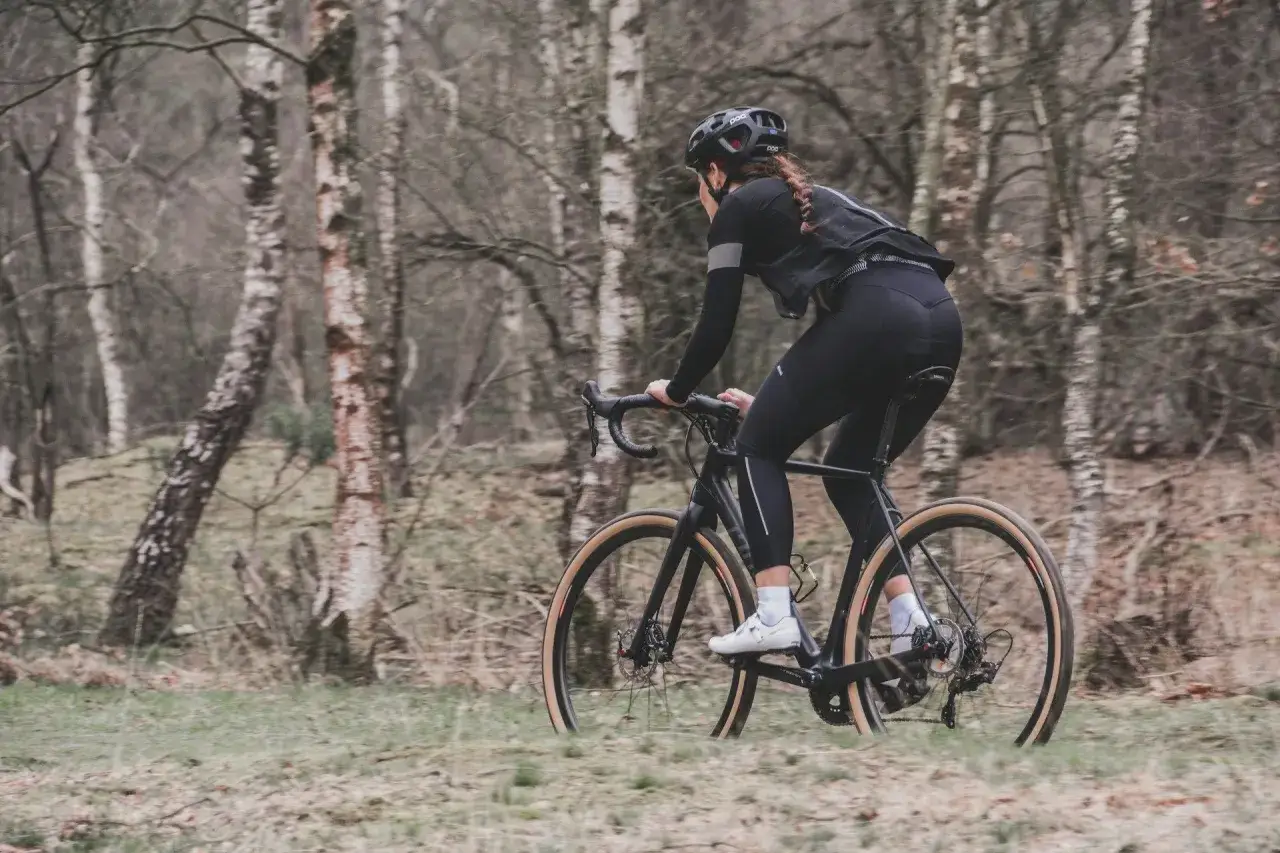 Kobieta w kasku i czarnym stroju jedzie na rowerze typu gravel przez las.
