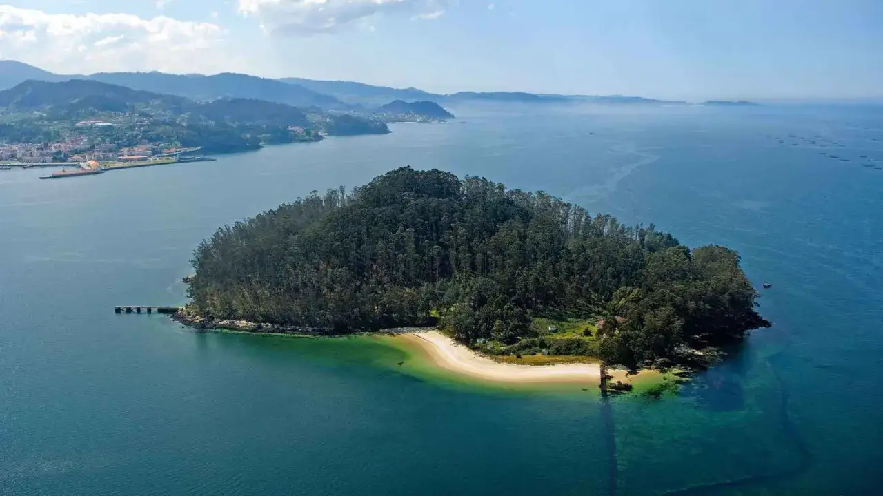 Isla de Tambo, una joya verde rodeada de aguas turquesas, con una playa dorada y un muelle. Al fondo, la costa gallega y el mar.