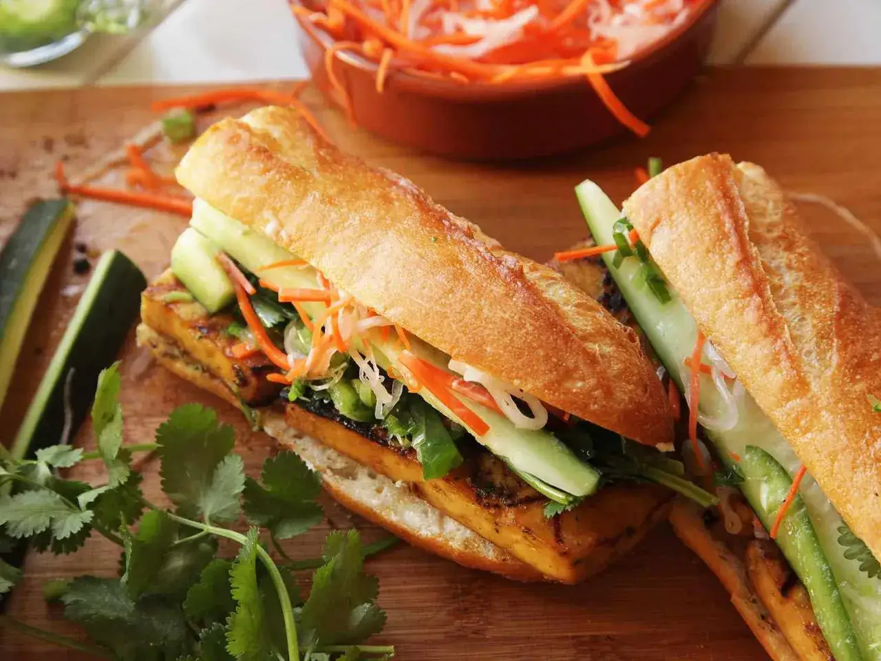 Två banh mi tofu-smörgåsar med grillad tofu, gurka, morötter och koriander på en träskärbräda.