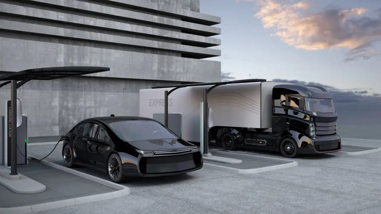 Un coche negro y un **camión del futuro** cargando en una estación eléctrica frente a un edificio de hormigón.