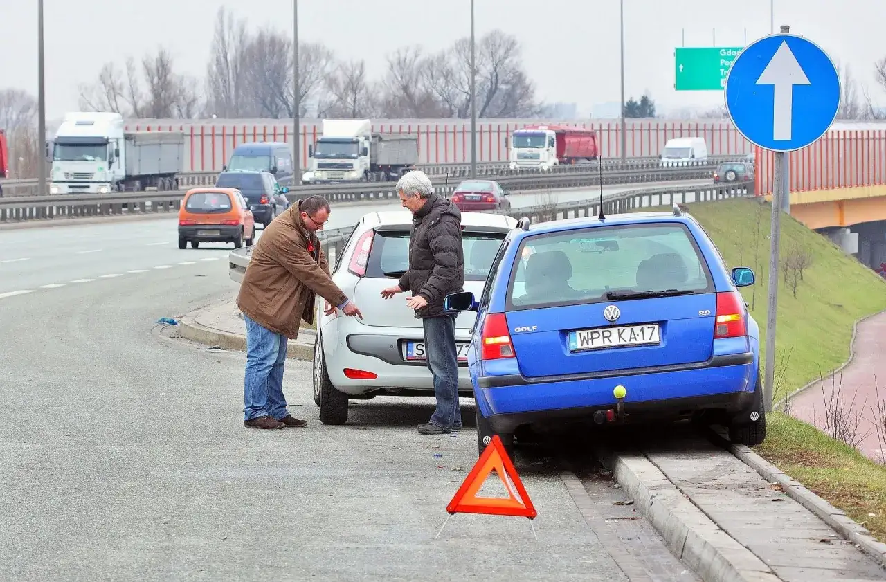 Dwóch mężczyzn dyskutuje przy samochodach po kolizji drogowej. Na drodze stoi trójkąt ostrzegawczy, a w tle widać ruchliwy wiadukt. Może czeka ich mandat.