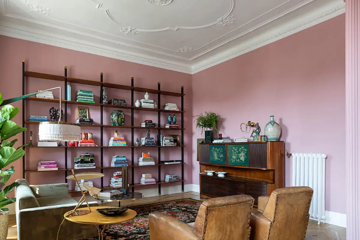 Accogliente salotto con pareti rosa cipria e mobili in legno scuro. Libreria piena di libri e oggetti decorativi, poltrone in pelle marrone e un tavolino giallo.