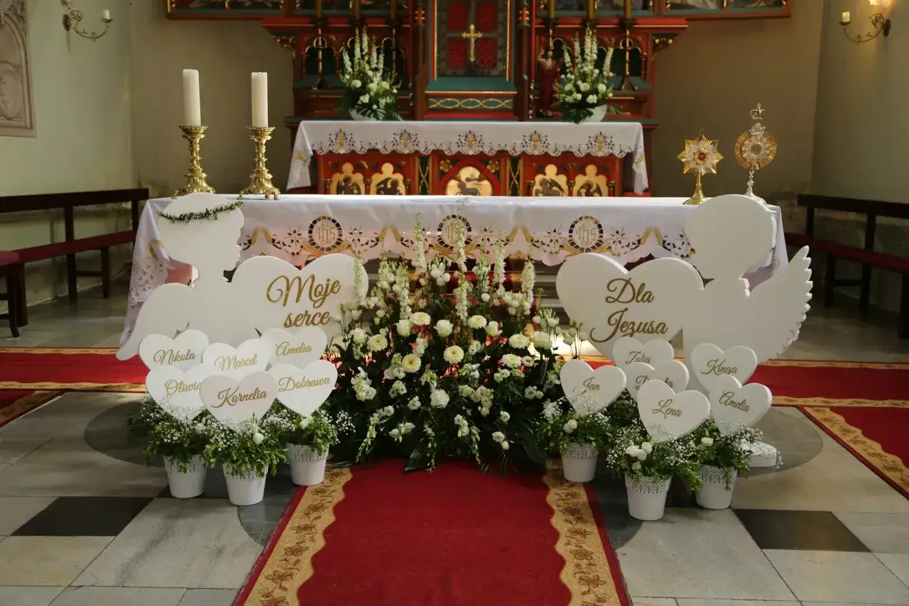 Dekoracje kościoła na komunię: aniołki, serca z imionami dzieci i kwiaty. Widać ołtarz i ławki. Koszty komunii w kościele to często wydatek na takie ozdoby.