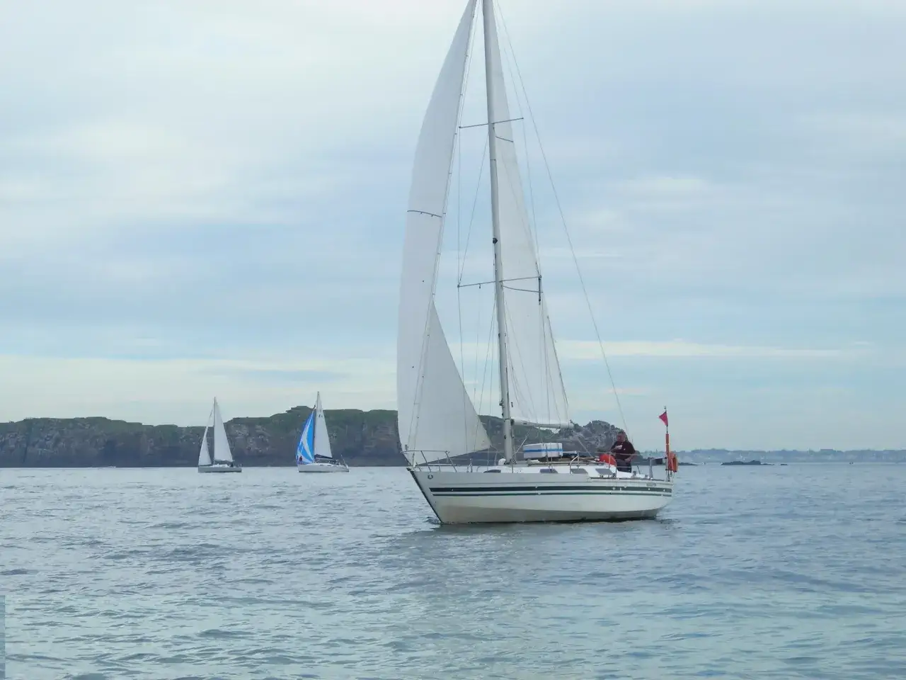 Un voilier Aquavit navigue sur une mer calme, suivi par d'autres voiliers. Le ciel est couvert.
