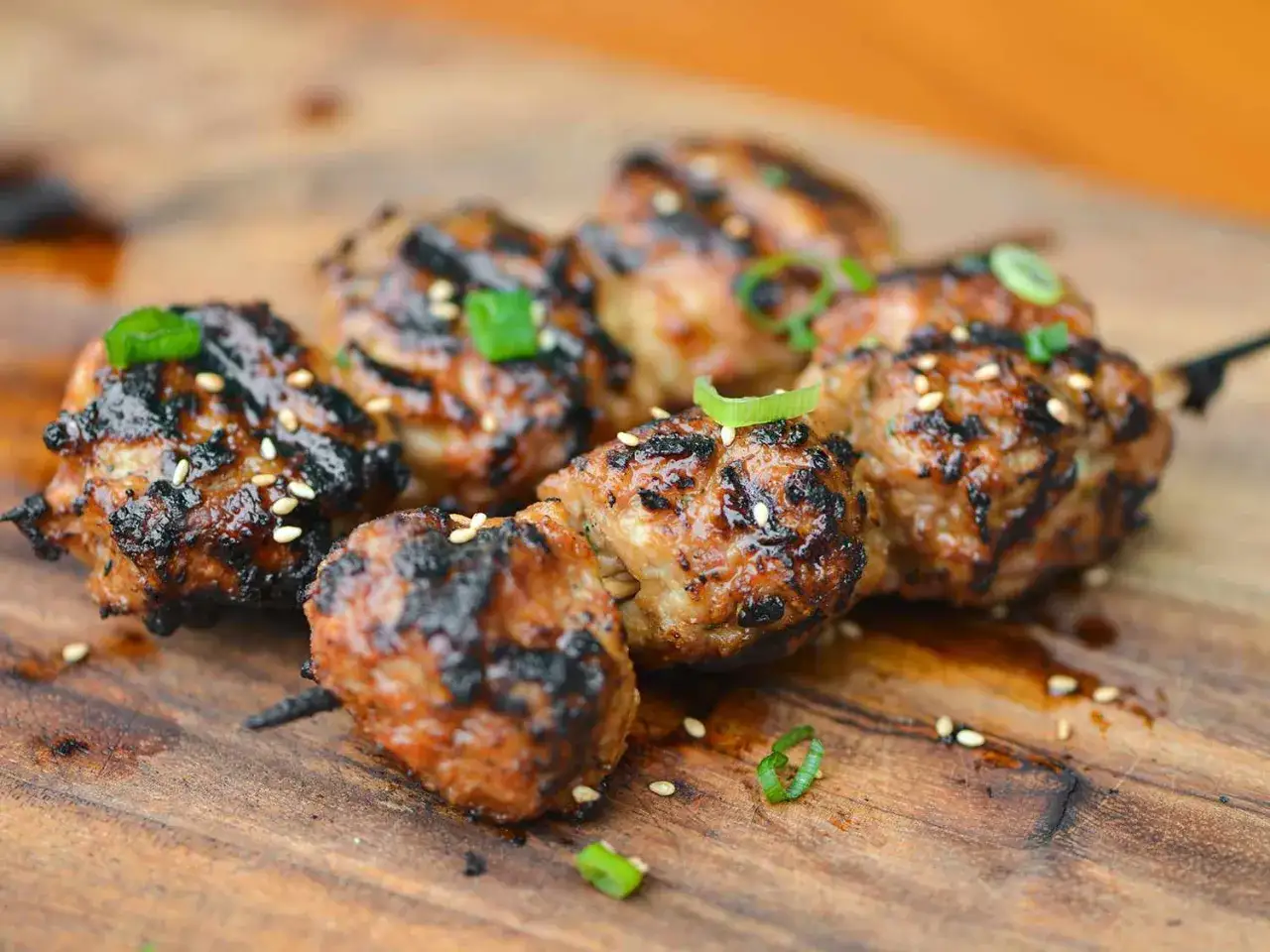 A skewer of grilled Japanese meatballs, glistening with sauce and sprinkled with sesame seeds and green onions, rests on a wooden board.