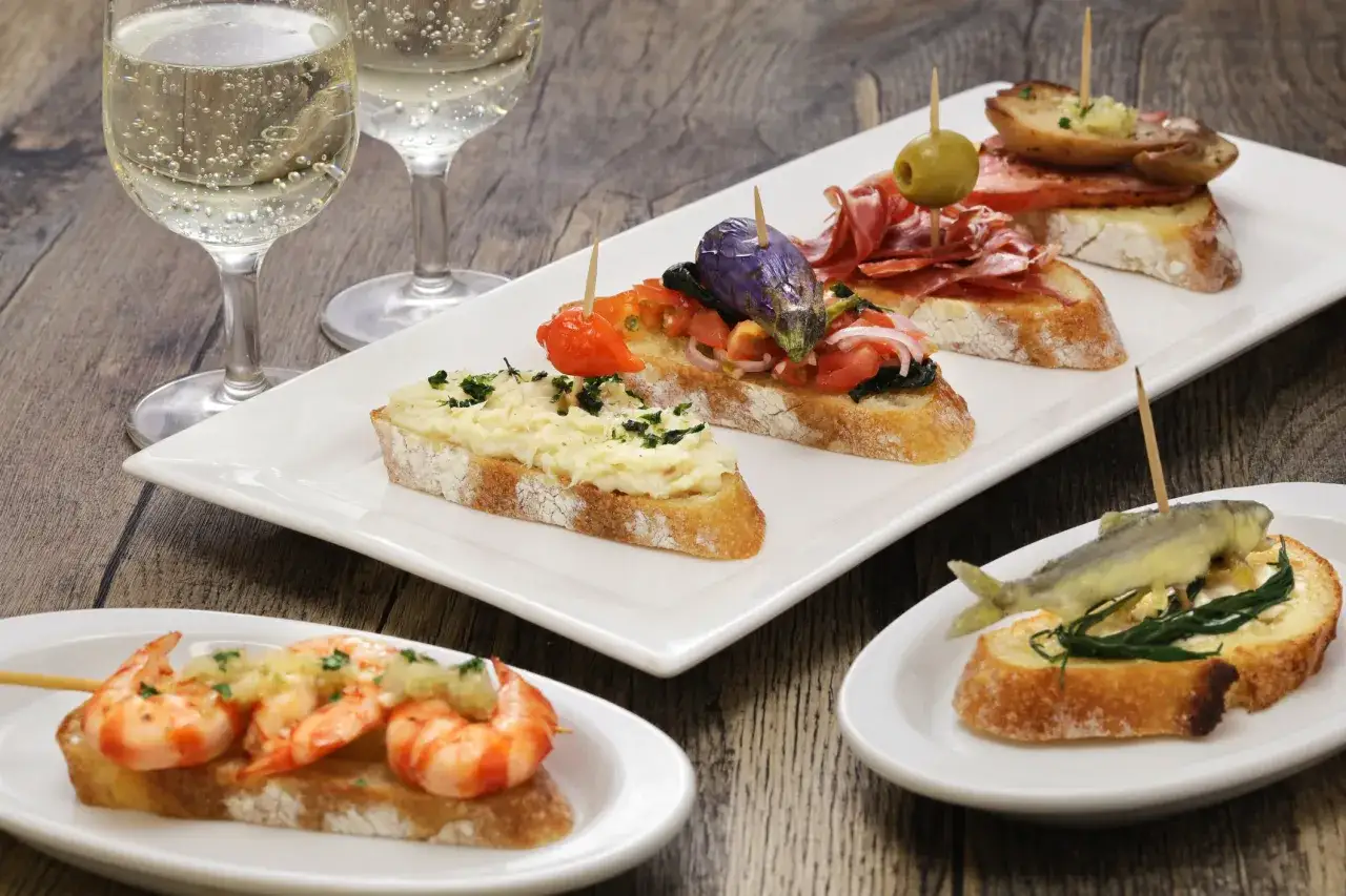 Pinchos variados, como gambas, pescado y jamón, acompañados de copas de vino espumoso, un festín que evoca el vino típico de Bilbao.