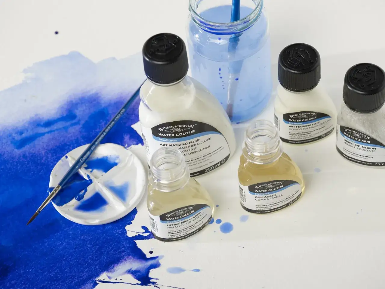 Pinceau sur palette avec des peintures bleues. Bouteilles de médiums pour aquarelle, dont la colle gomme arabique, sur fond blanc.