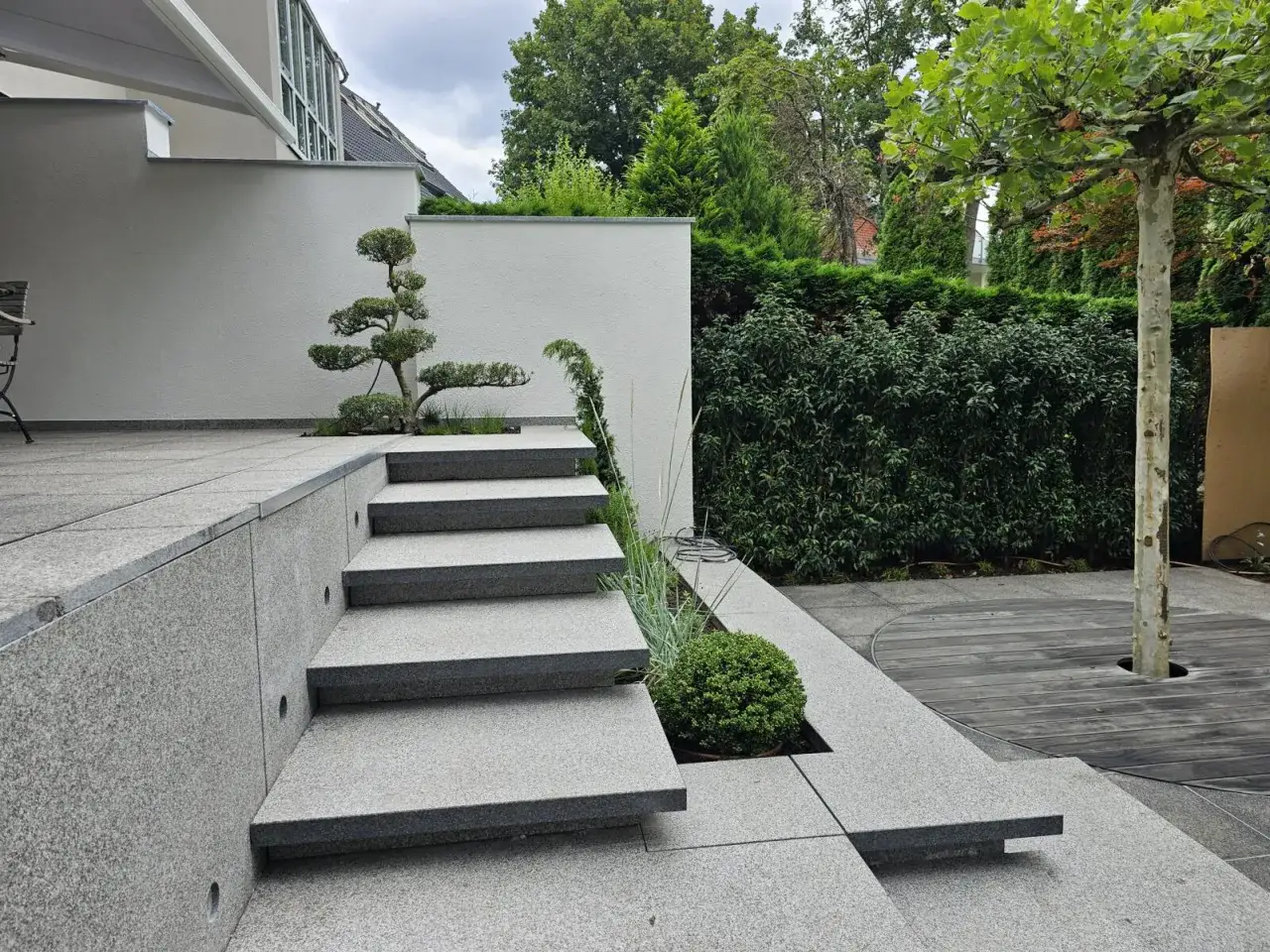 Moderne Ideen f&uuml;r den Hauseingang: Eine elegante Steintreppe f&uuml;hrt zu einer Terrasse, umgeben von gepflegten Gr&uuml;nfl&auml;chen und einem Baum.