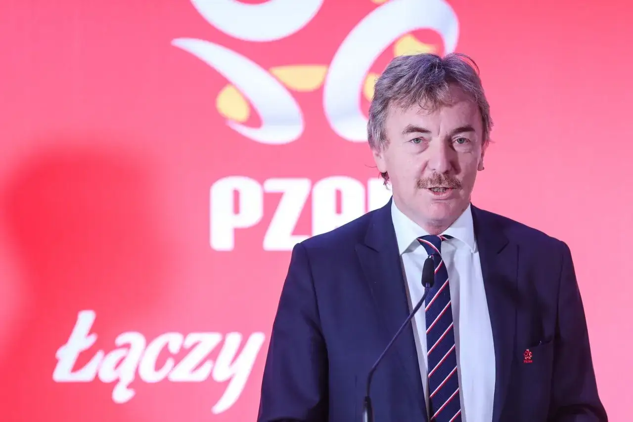 Prezes PZPN, Cezary Kulesza, w garniturze i krawacie przemawia na tle czerwonej ścianki z logo związku.
