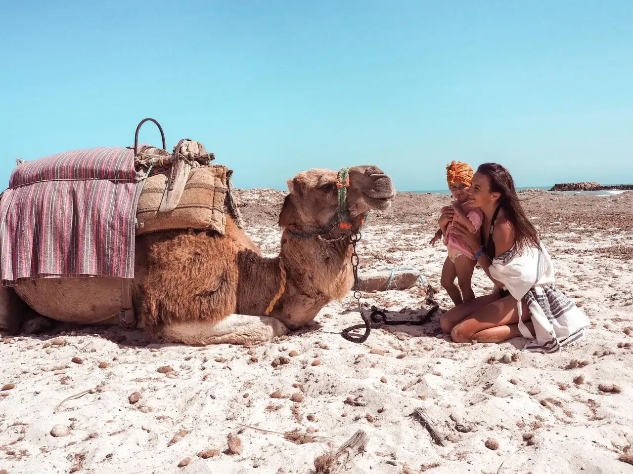 Djerba na co uważać? Kobieta z dzieckiem na plaży obok wielbłąda.