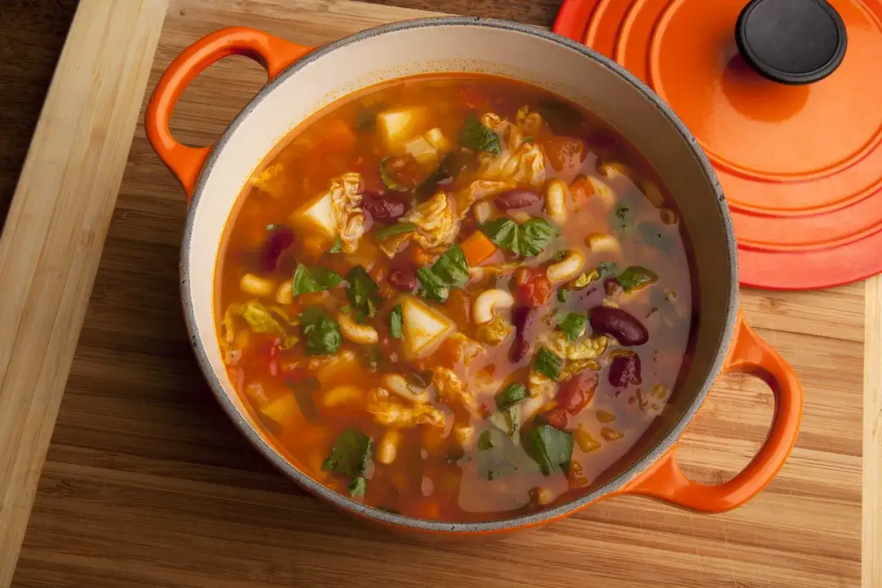 Sopa minestrone humeante en olla naranja, llena de verduras, frijoles y hierbas frescas.