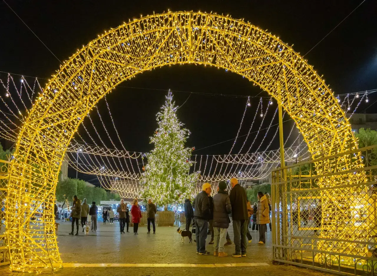Un arco di luci dorate illumina la piazza di Urbino Natale, con un albero addobbato e persone che passeggiano.