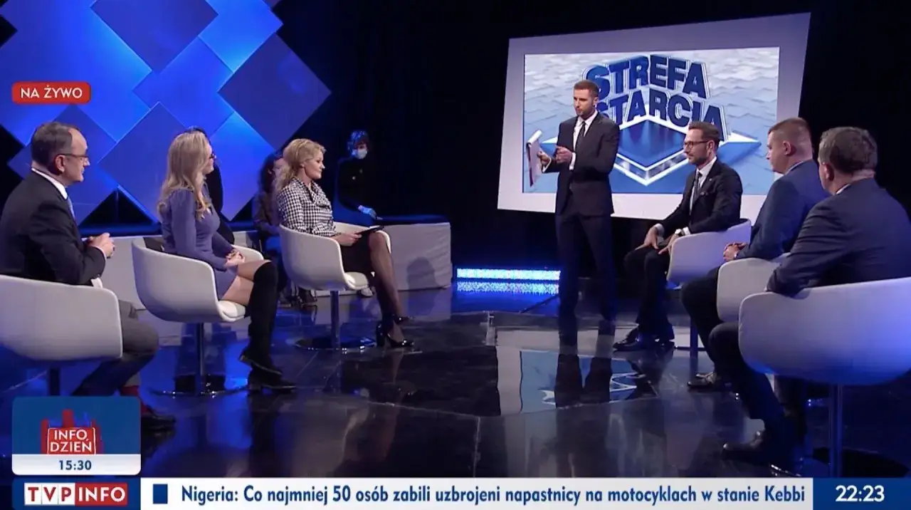 W tle wizji prowadzący dyskutuje z gośćmi siedzącymi w studiu.