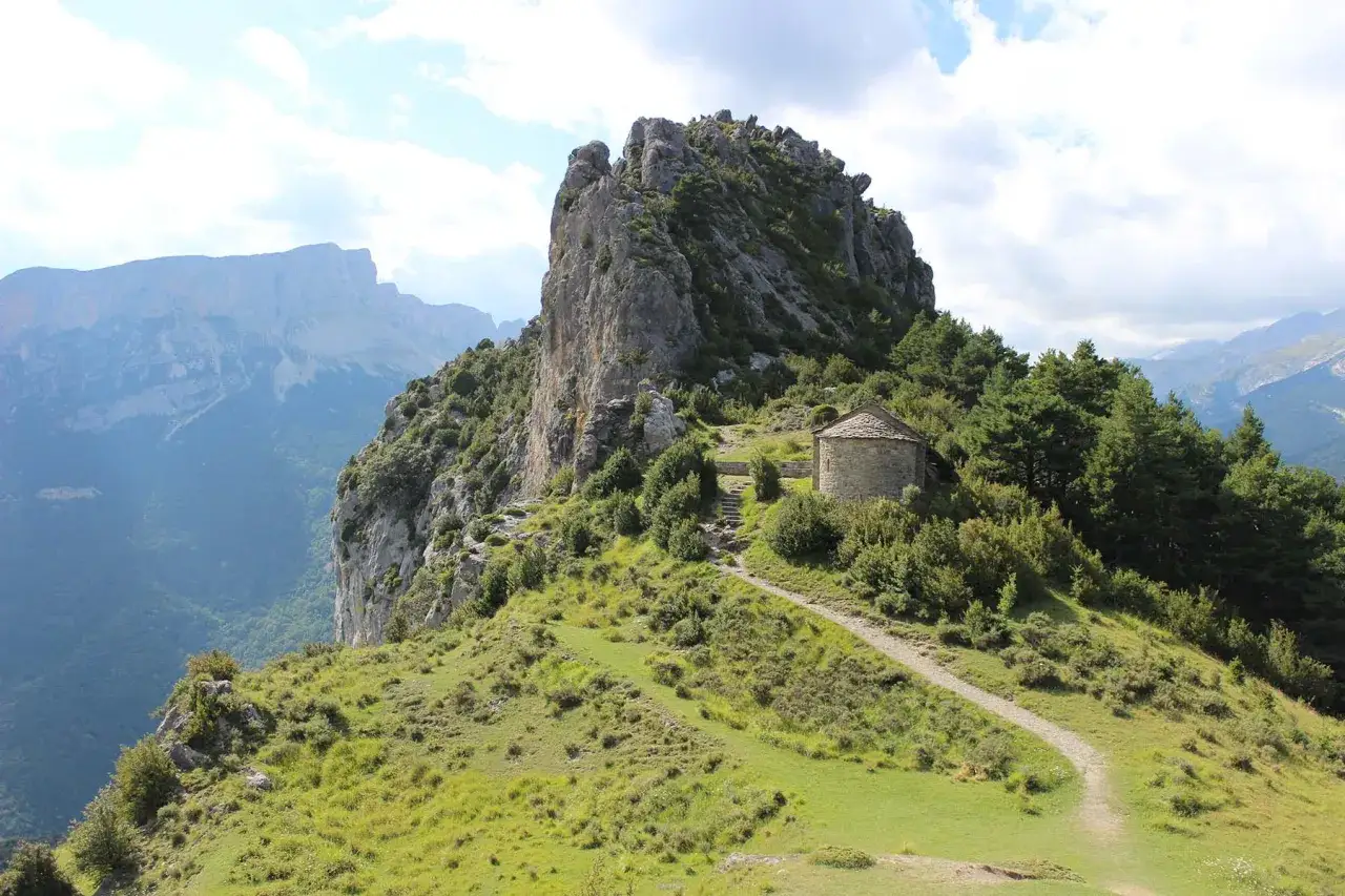 Pequeña ermita de piedra en el **valle de Chistau**, rodeada de vegetación y con imponentes montañas al fondo.