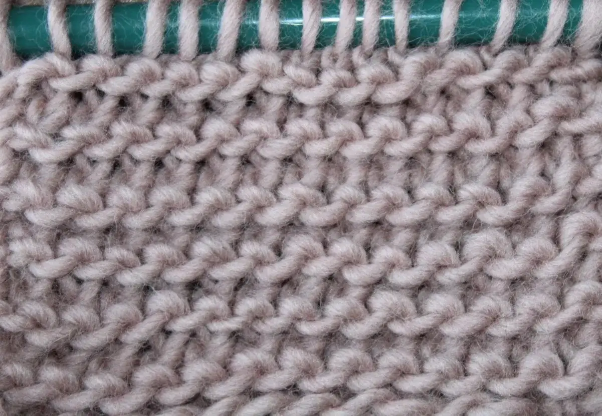 Tejido de lana color beige con punto Santa Clara, mostrando la textura de las hebras y las agujas verdes al inicio.