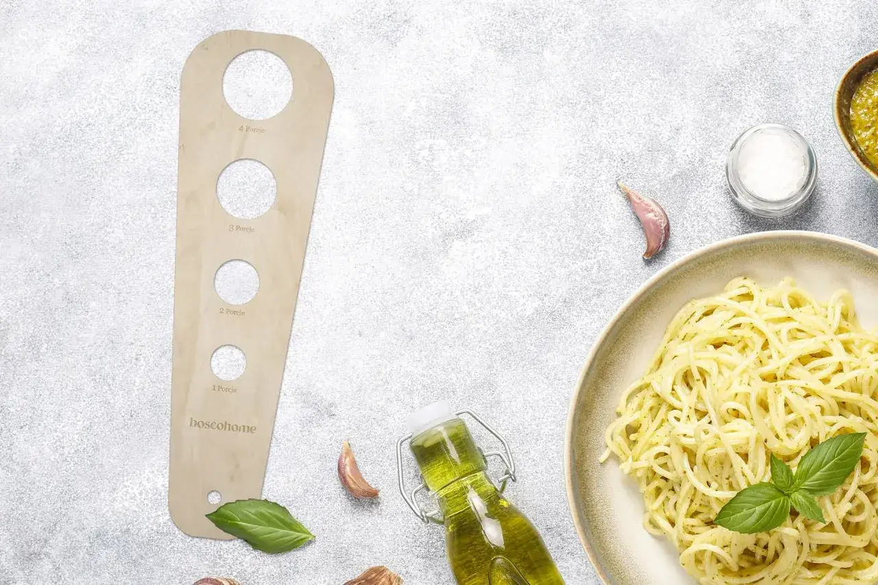 Drewniany przyrząd do odmierzania, ile makaronu na porcję, obok talerza spaghetti, butelki oliwy i czosnku.