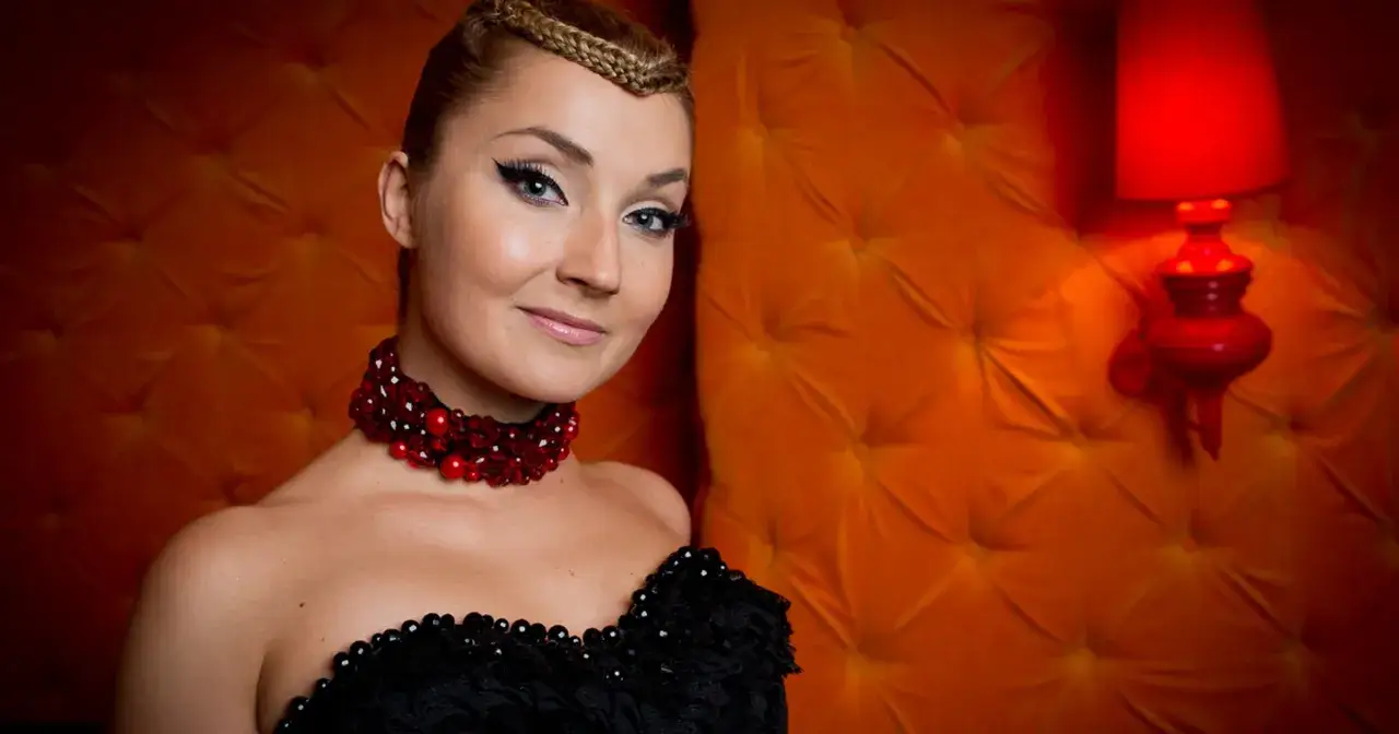 Cleo w czarnej sukni i czerwonej biżuterii, z warkoczem na głowie, pozuje na tle pomarańczowej, pikowanej ściany.