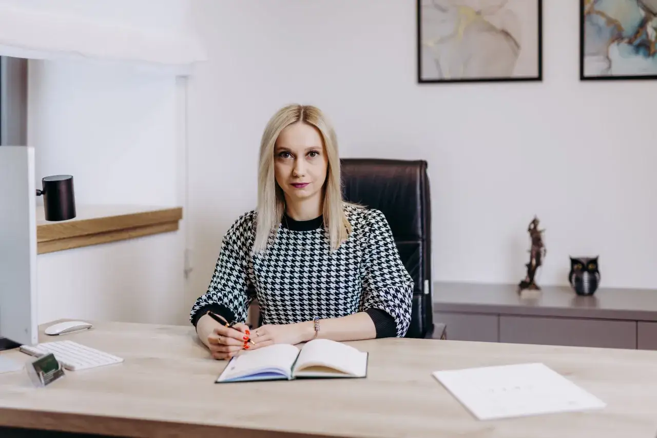 Kobieta w czarno-białej sukience siedzi przy biurku, pisząc w notatniku.