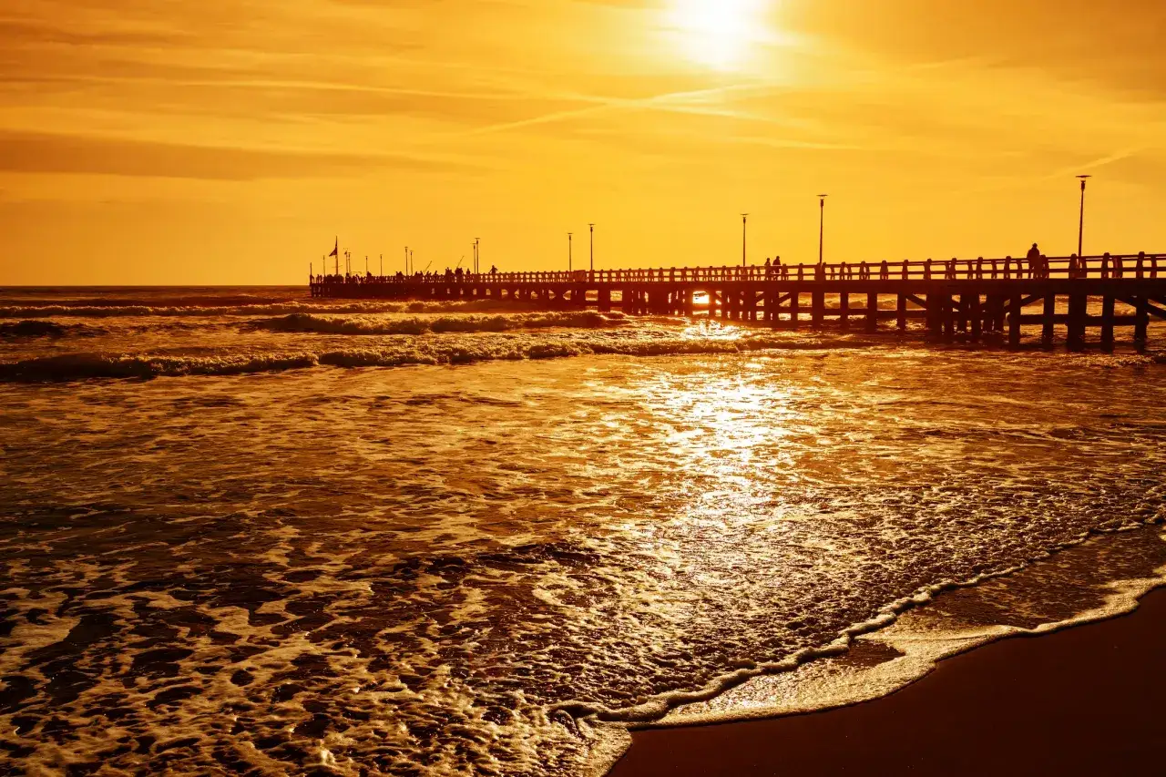 Tramonto dorato sul pontile di Forte dei Marmi, Italia. Le onde lambiscono la riva mentre le sagome delle persone passeggiano.