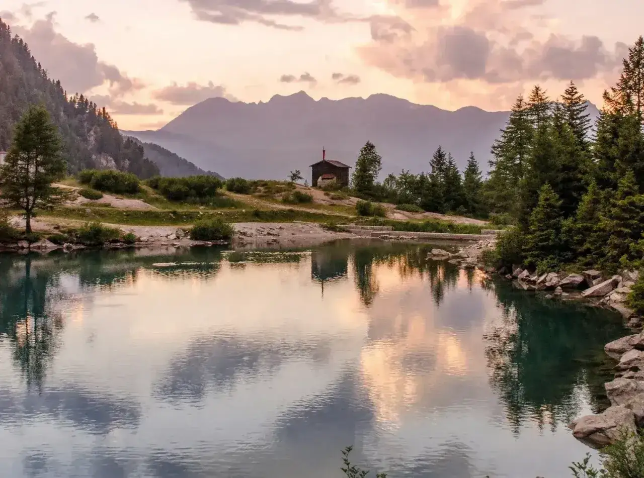 Riflessi di un tramonto su un lago alpino, con un piccolo rifugio in legno in lontananza. Un luogo incantevole dove scoprire cosa vedere, magari attraversando un ponte di legno.