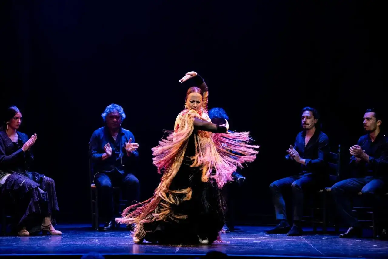 Bailarina de flamenco en pleno movimiento, con un mantón de flecos vibrantes, rodeada de músicos y público aplaudiendo en conciertos flamenco zaragoza.