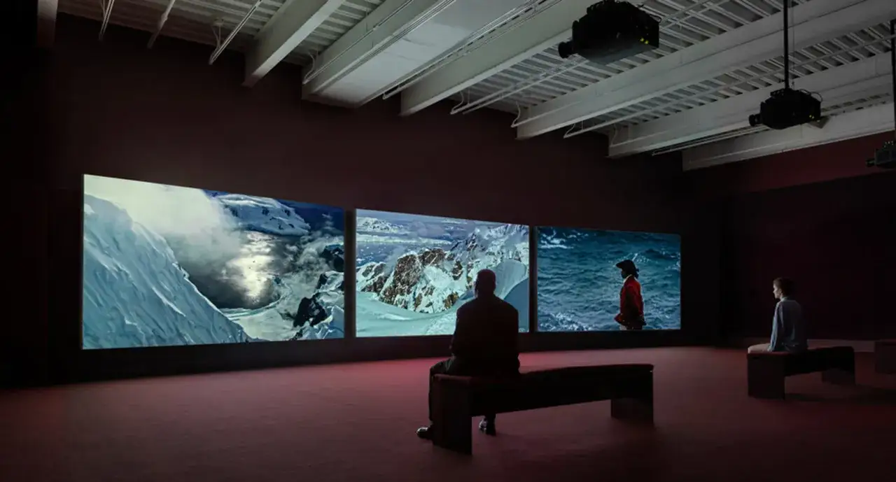A triptych screen displays John Akomfrah art: icy landscapes and a figure in red against the sea. Two viewers sit in contemplation.