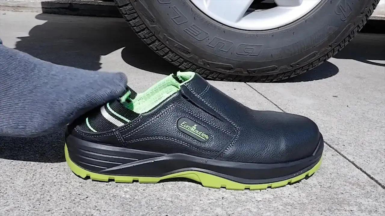 Un pied en chaussette grise s'apprête à enfiler une chaussure de sécurité noire et verte, prête pour conduire en claquette.